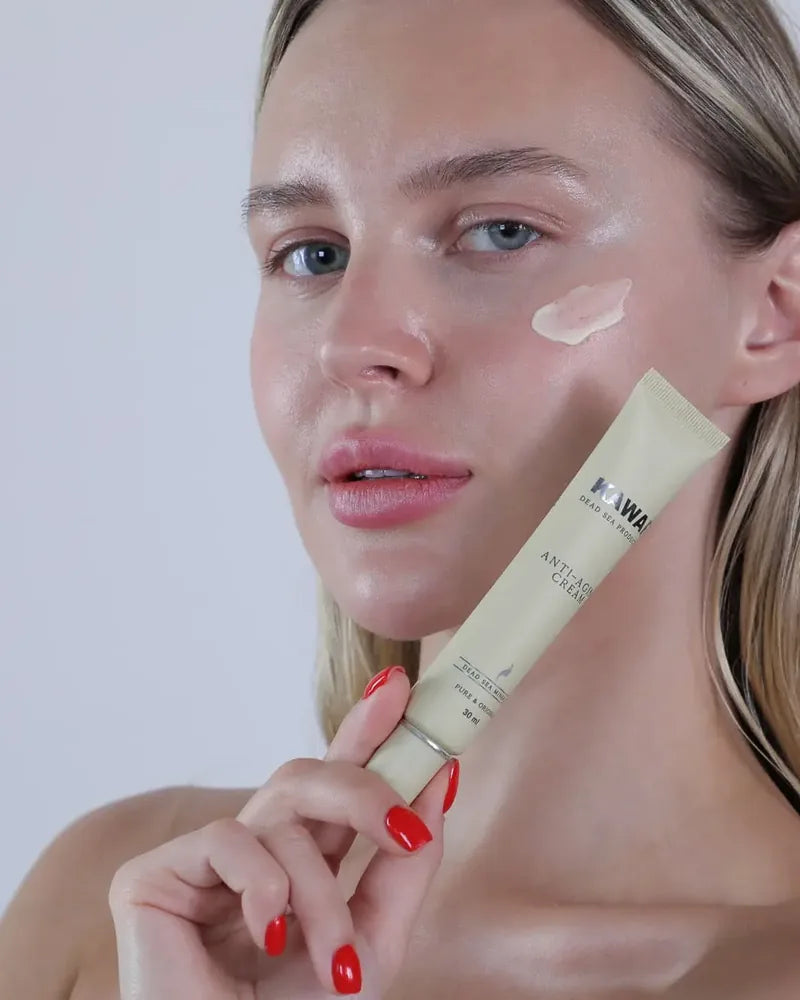 A close-up of a woman holding a beige tube of Kawar Dead Sea Products Anti-Aging Cream while applying a dollop of the cream to her cheek. She has blonde hair, blue eyes, and red manicured nails, set against a clean, light gray background.